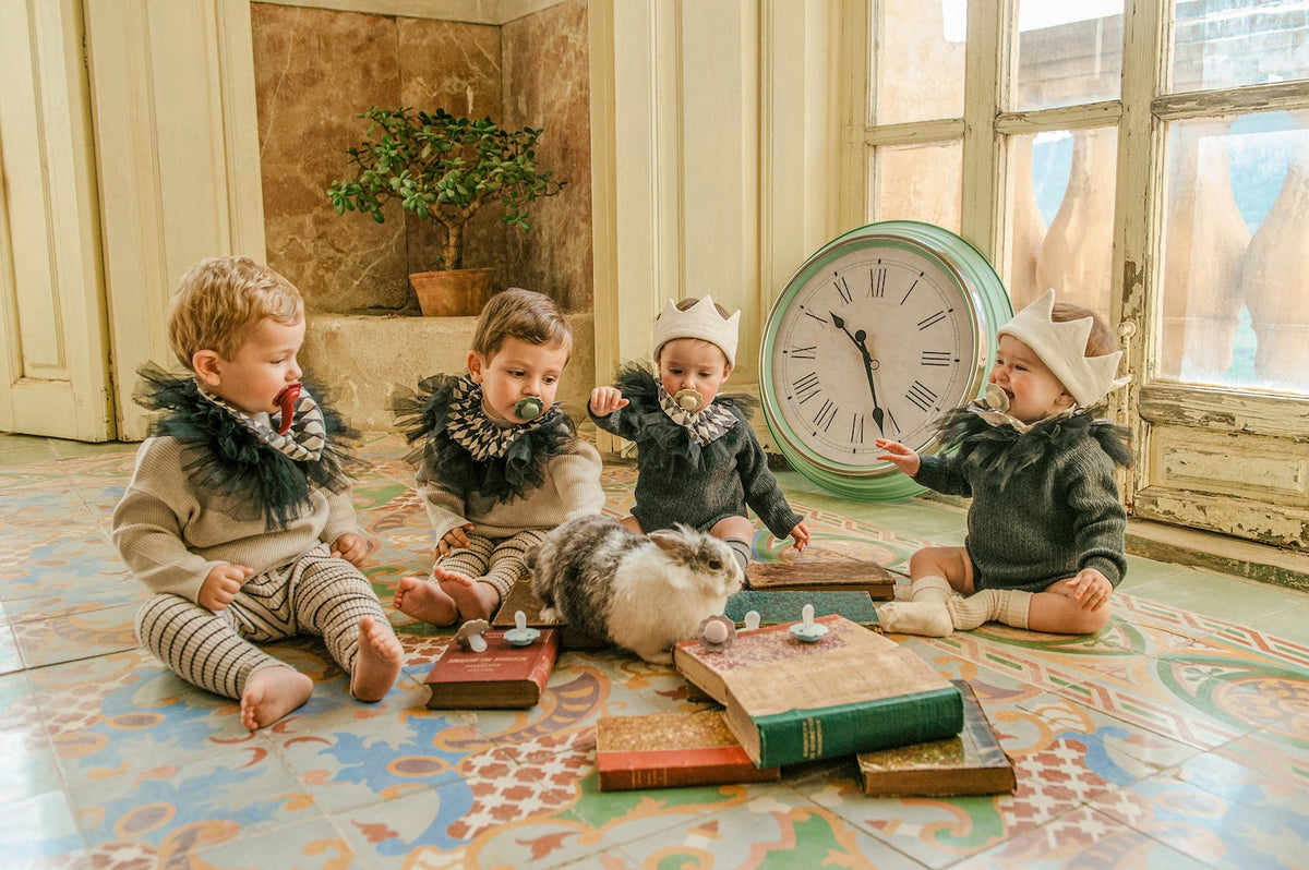Four babies in ruffled collars sitting on a tiled floor with Suavinex Wonder soothers